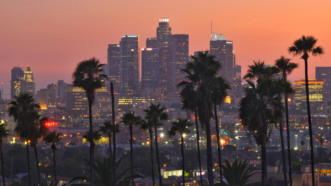 Panoramic view of the California skyline at sunset, featuring modern high-rise buildings silhouetted against a vibrant orange and pink sky.